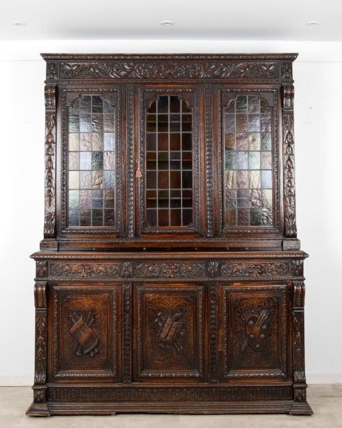 fig 1. A large late Victorian dark carved oak bookcase featuring three upper glazed doors with coloured leaded glass panels above a lower cupboard base with relief carved emblems.