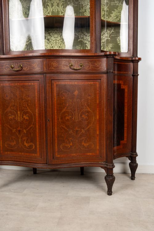 fig 2. An Edwardian mahogany display cabinet featuring satinwood marquetry inlay, glazed upper doors with green fabric lining, and turned legs.