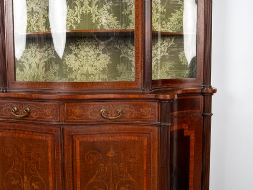 fig 3. An Edwardian mahogany display cabinet featuring satinwood marquetry inlay, glazed upper doors with green fabric lining, and turned legs.