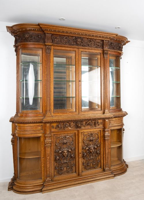 fig 2. A large Victorian carved oak display cabinet featuring curved glass sides, intricately carved mask motifs on lower door panels, and glass shelving.