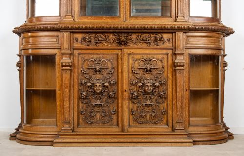 fig 3. A large Victorian carved oak display cabinet featuring curved glass sides, intricately carved mask motifs on lower door panels, and glass shelving.