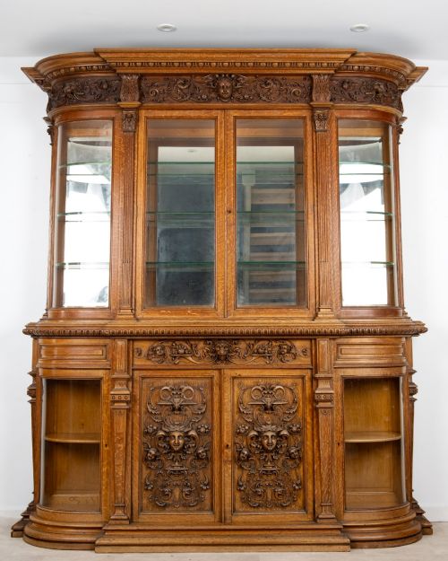 fig 4. A large Victorian carved oak display cabinet featuring curved glass sides, intricately carved mask motifs on lower door panels, and glass shelving.