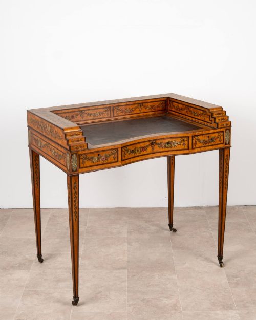 fig 1. A warm gold satinwood Carlton House writing desk featuring hand-painted floral swags, a dark leather writing surface, and tapered legs on brass castors.