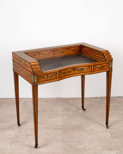 fig 2. A warm gold satinwood Carlton House writing desk featuring hand-painted floral swags, a dark leather writing surface, and tapered legs on brass castors.