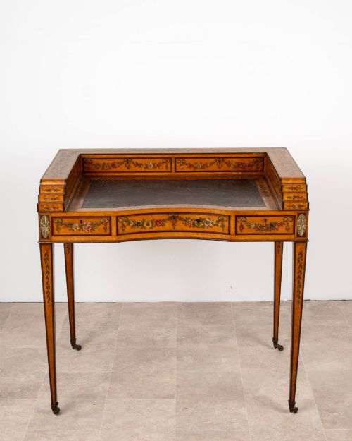 fig 3. A warm gold satinwood Carlton House writing desk featuring hand-painted floral swags, a dark leather writing surface, and tapered legs on brass castors.