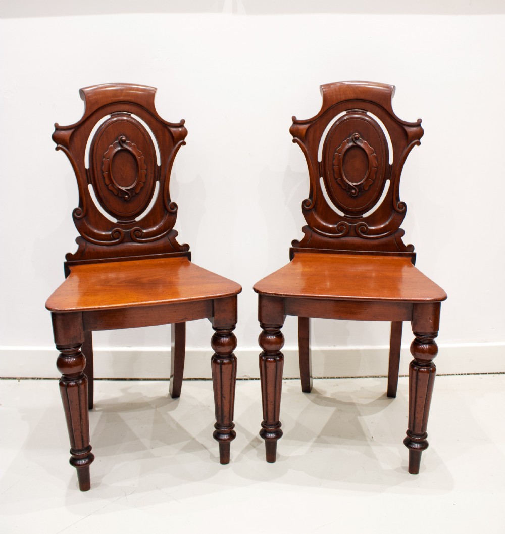 A pair of 19th-century mahogany hall chairs with carved oval cartouche backs, flat timber seats, and turned front legs. View II