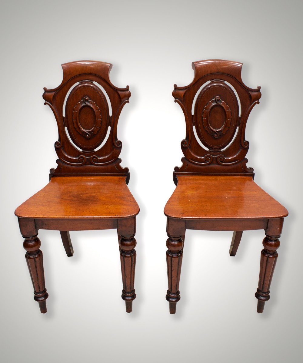 A pair of 19th-century mahogany hall chairs with carved oval cartouche backs, flat timber seats, and turned front legs.