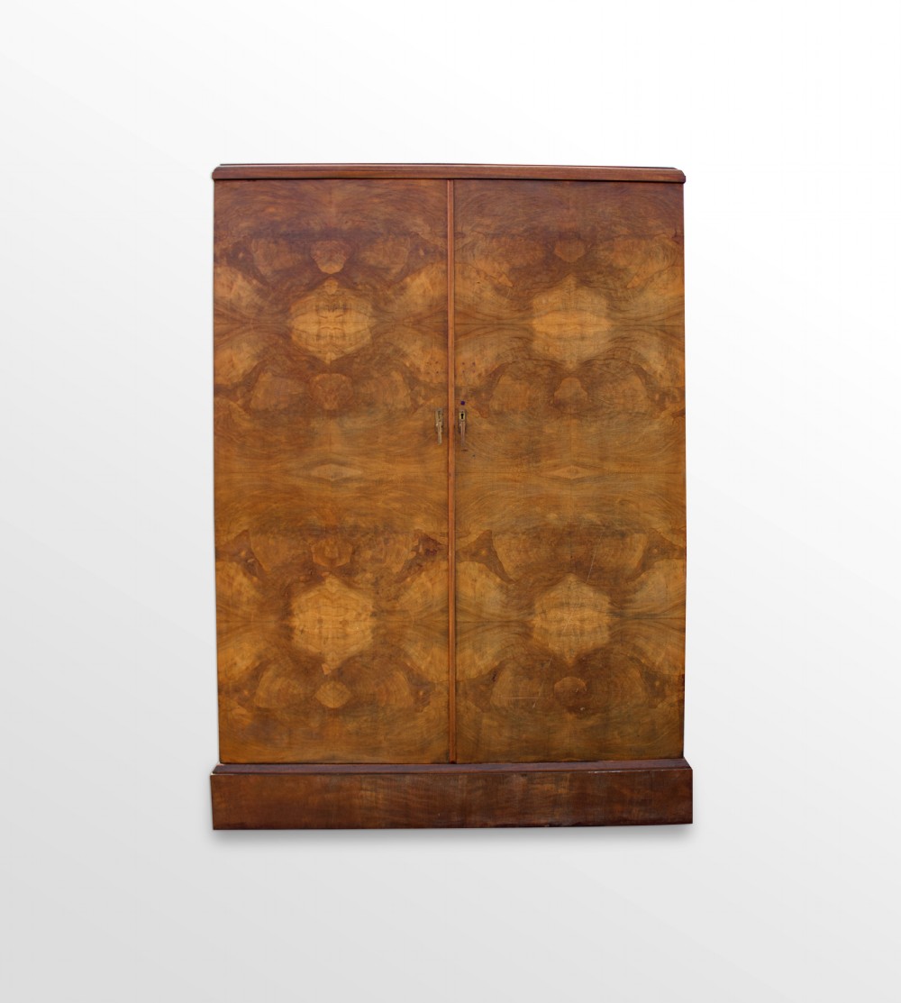 An Art Deco two-door compactum wardrobe crafted from book-matched burr walnut veneer with central keyholes and a solid plinth base.