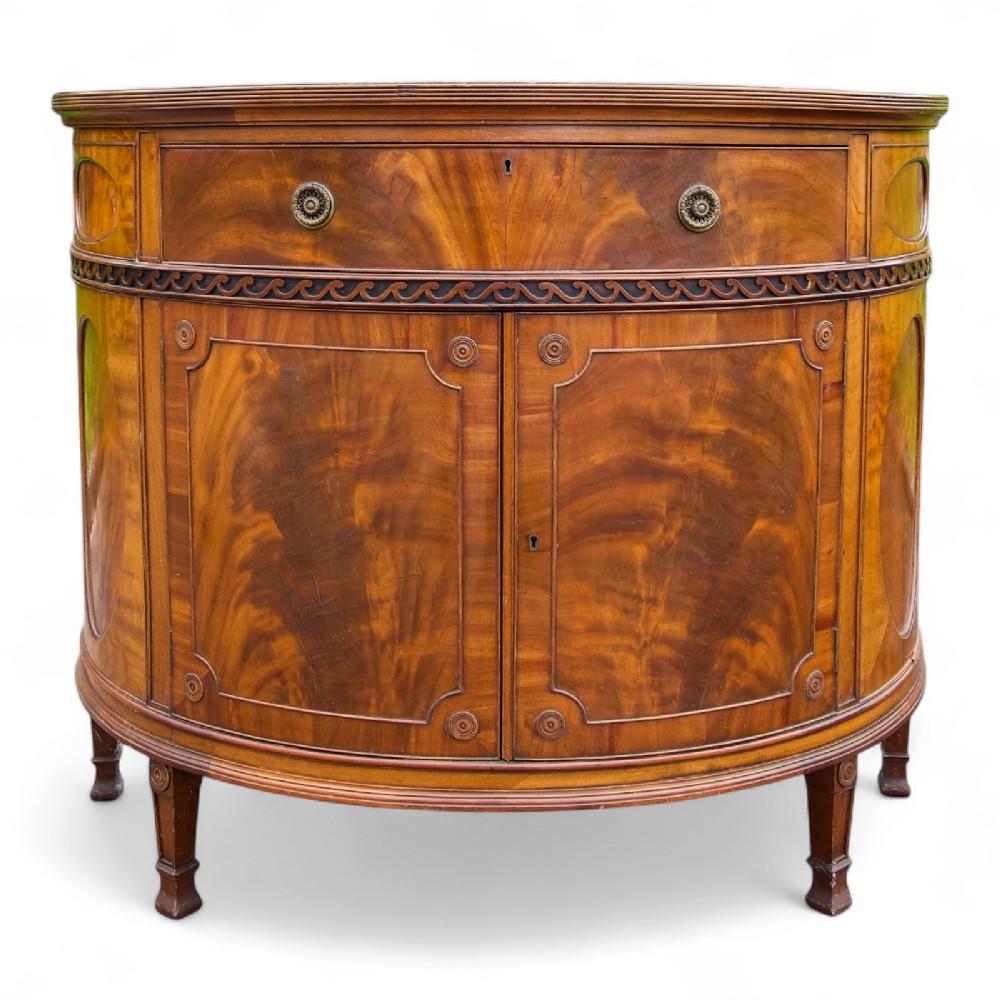 An antique flamed mahogany demi lune commode featuring a curved front, single top drawer, two cabinet doors, and circular brass ring handles.
