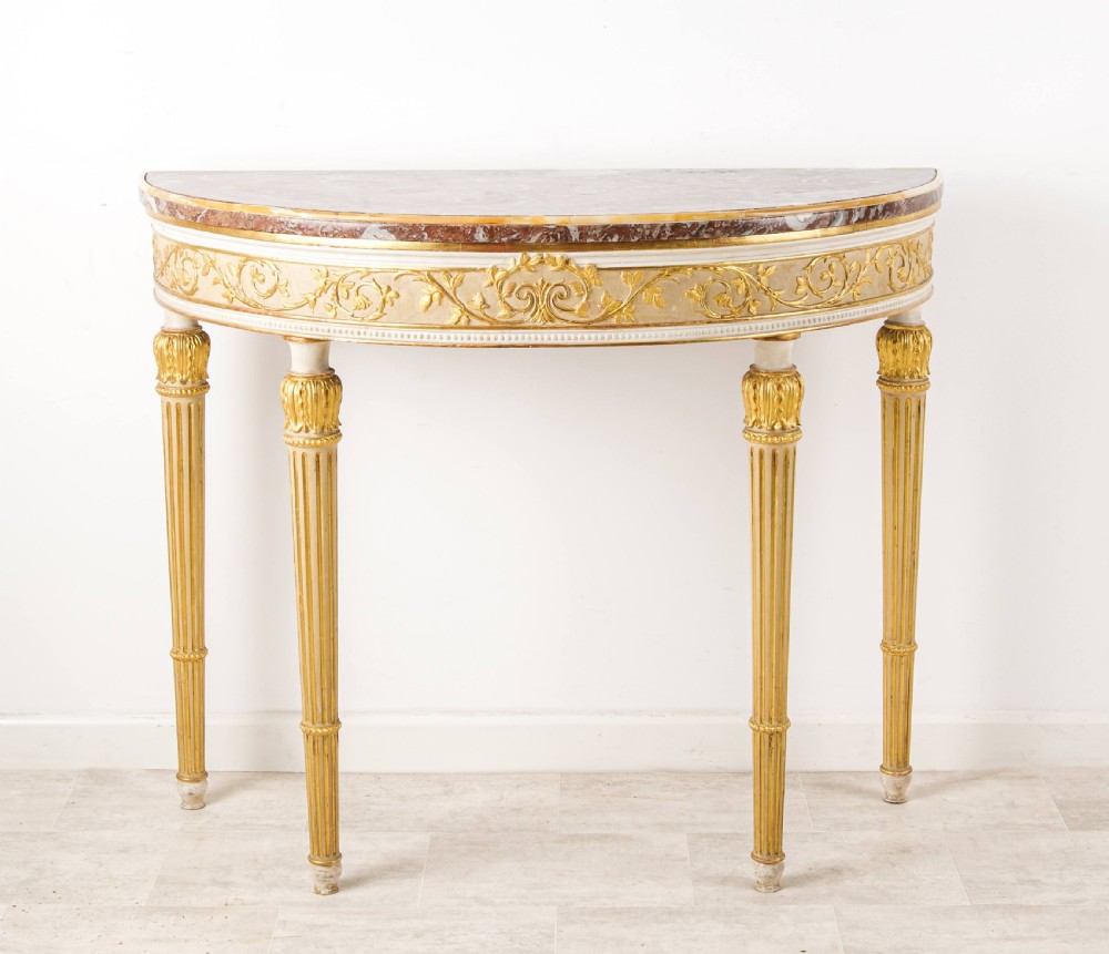 A Georgian style demi-lune console table featuring a rouge marble top, gilded foliate carved apron, and four fluted tapered legs.