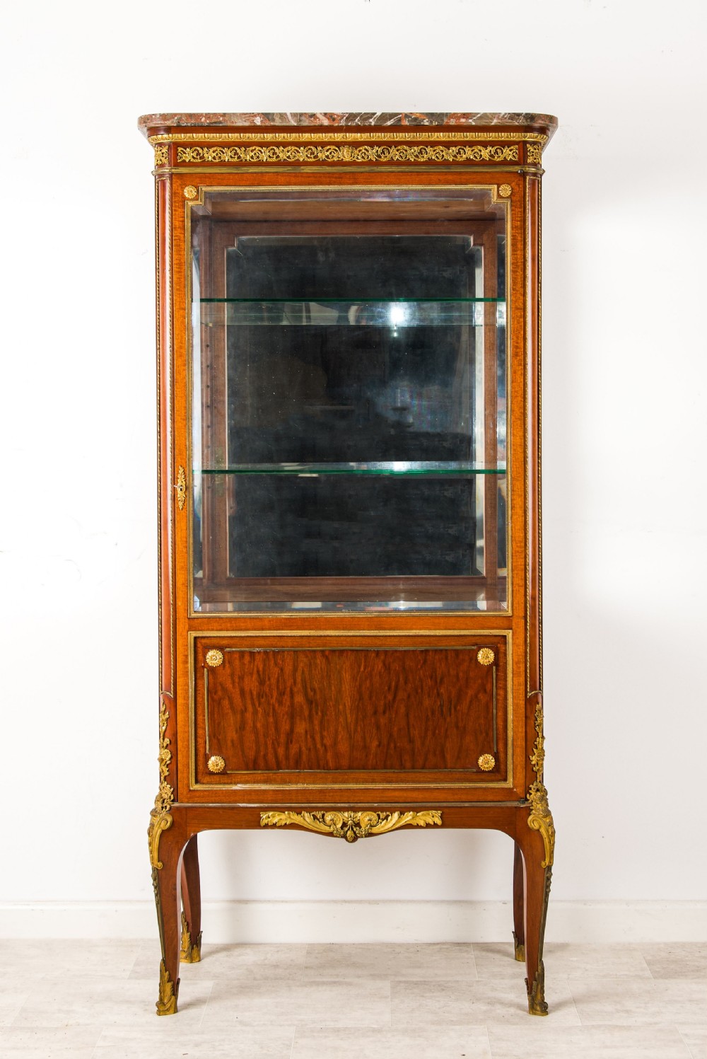 A French mahogany vitrine cabinet featuring glass shelves, ornate gilt-bronze mounts, cabriole legs, a mirrored back interior, and a marble top.