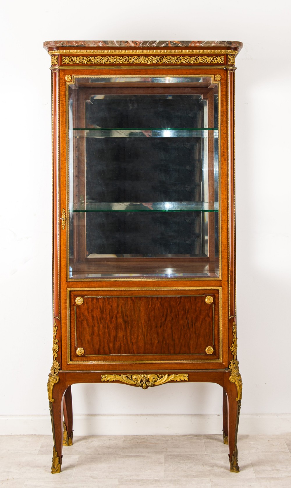 A French mahogany vitrine cabinet featuring glass shelves, ornate gilt-bronze mounts, cabriole legs, a mirrored back interior, and a marble top. View II