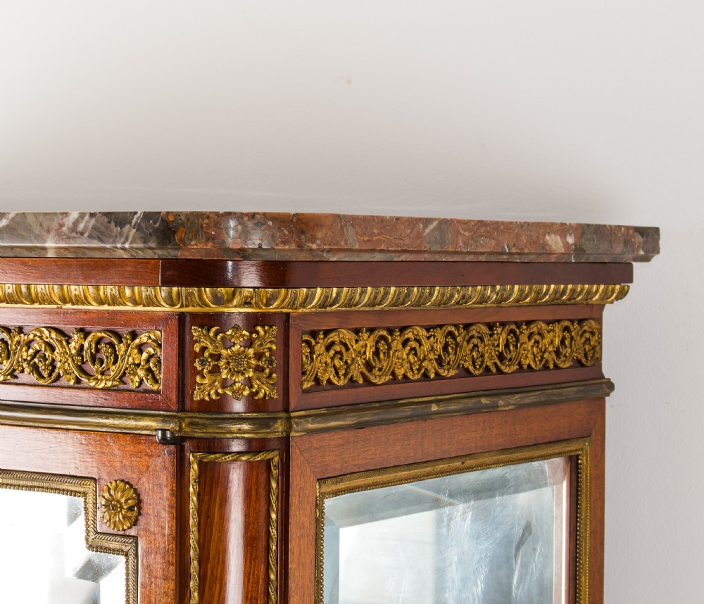 A French mahogany vitrine cabinet featuring glass shelves, ornate gilt-bronze mounts, cabriole legs, a mirrored back interior, and a marble top. View V