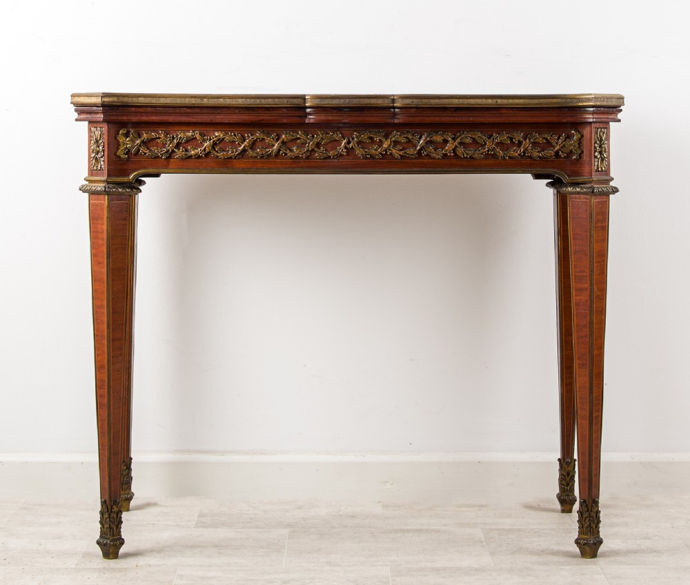 A French Louis XVI style games table featuring a parquetry fold-over top, foliage ormolu frieze, and square tapered legs with gilt mounts.