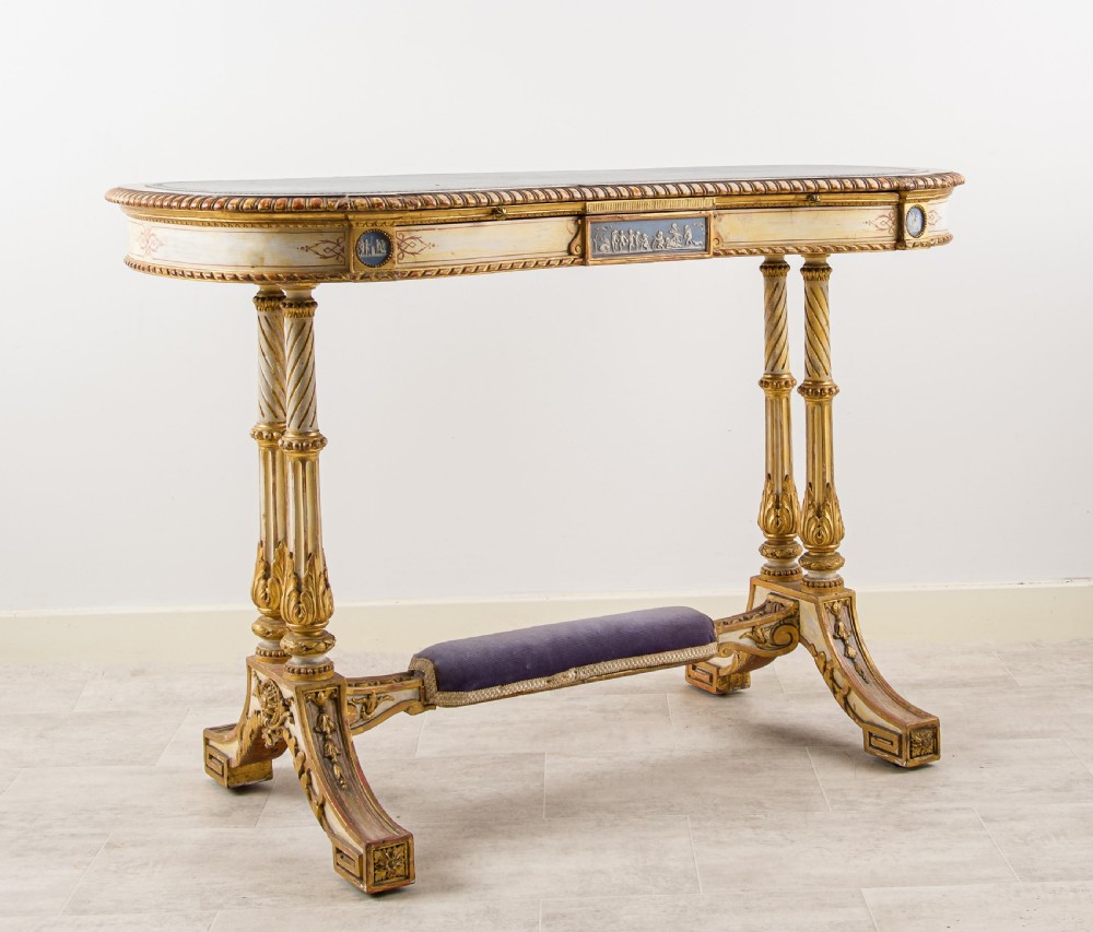 A 19th-century Neoclassical kidney-shaped writing table decorated in cream paint and gold leaf, featuring fluted legs, blue jasperware plaques, and a purple fabric-covered stretcher footrest.