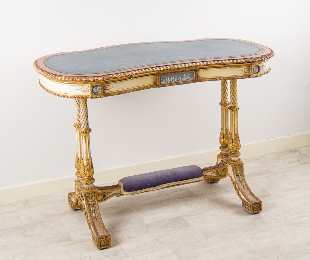 A 19th-century Neoclassical kidney-shaped writing table decorated in cream paint and gold leaf, featuring fluted legs, blue jasperware plaques, and a purple fabric-covered stretcher footrest. View II