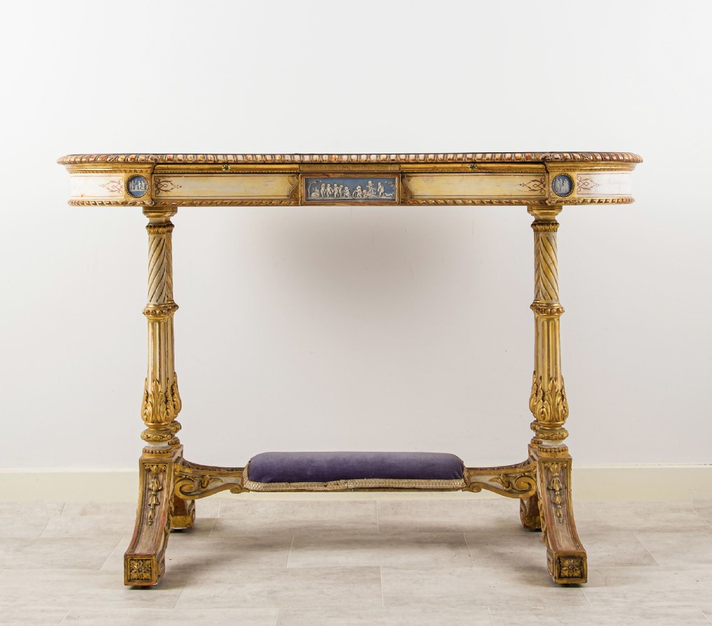 A 19th-century Neoclassical kidney-shaped writing table decorated in cream paint and gold leaf, featuring fluted legs, blue jasperware plaques, and a purple fabric-covered stretcher footrest. View VII