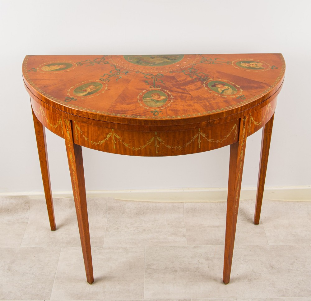 An antique satinwood demi-lune games table featuring hand-painted floral medallions and swags across the fold-over top and frieze.