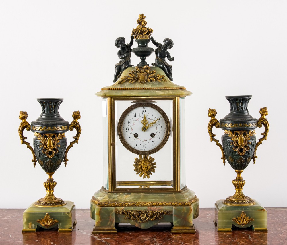 A French three-piece garniture set featuring a central glazed onyx clock topped with dark bronze cherub figures, flanked by two matching side urns on green onyx bases.