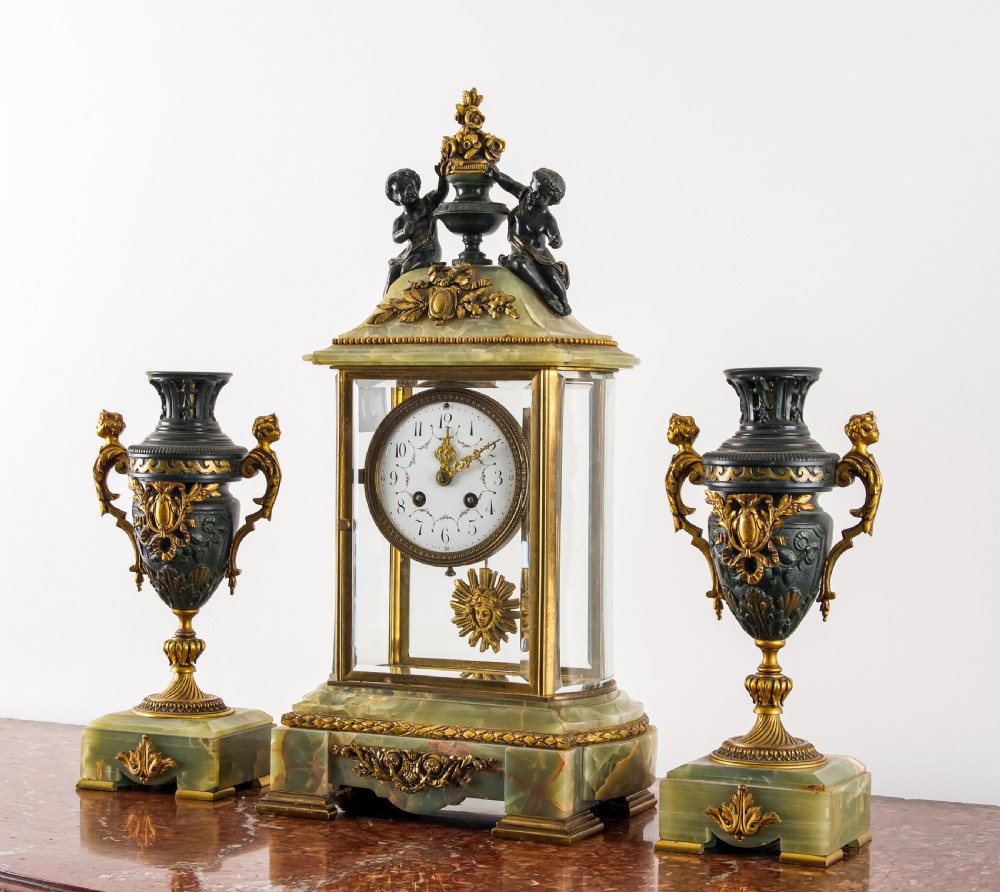 A French three-piece garniture set featuring a central glazed onyx clock topped with dark bronze cherub figures, flanked by two matching side urns on green onyx bases. View II