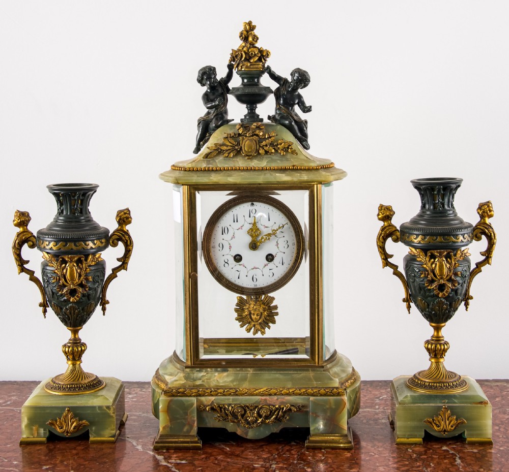 A French three-piece garniture set featuring a central glazed onyx clock topped with dark bronze cherub figures, flanked by two matching side urns on green onyx bases. View III