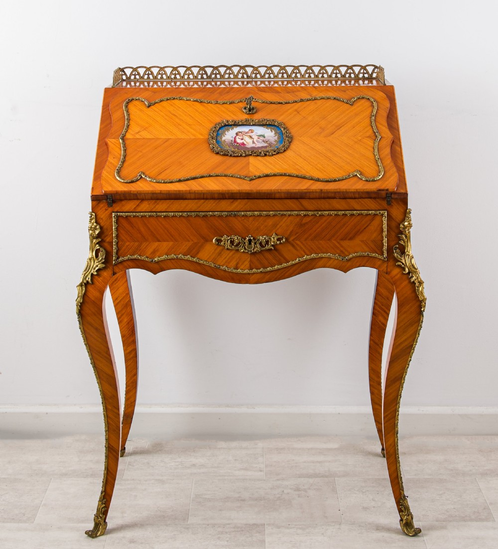 An antique French Louis XV style kingwood writing bureau with a slant-front lid featuring a central painted porcelain plaque, brass top gallery, cabriole legs, and gilded ormolu mounts.