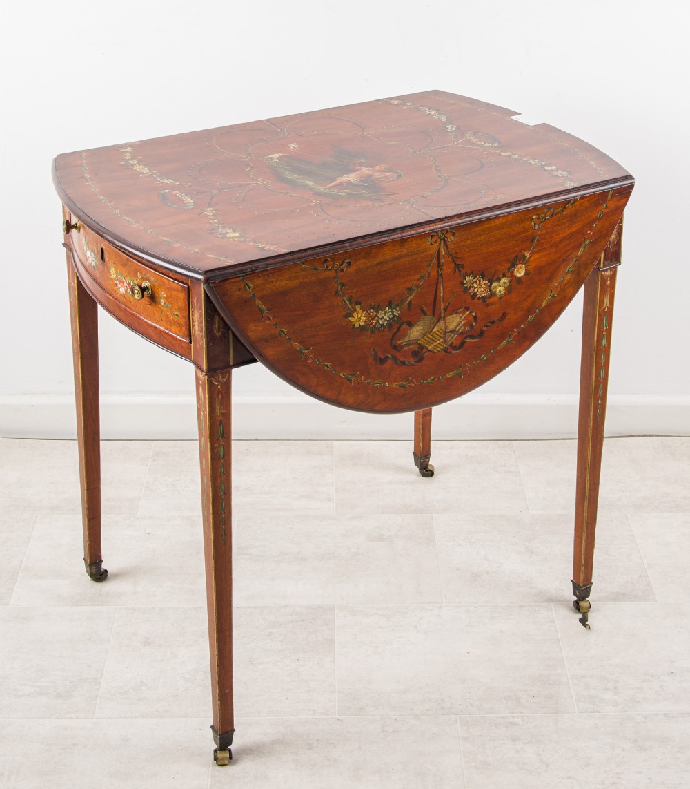 An antique mahogany Pembroke table featuring hand-painted floral swags, tapered legs with brass castors, and two hinged drop leaves.