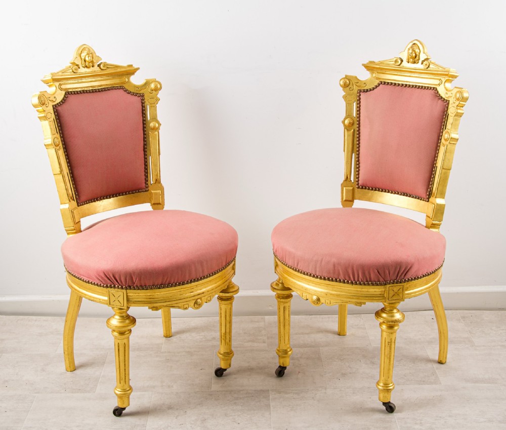 A pair of antique French giltwood salon chairs featuring carved cresting, pink velvet upholstered seats and backs, and fluted front legs on castors.