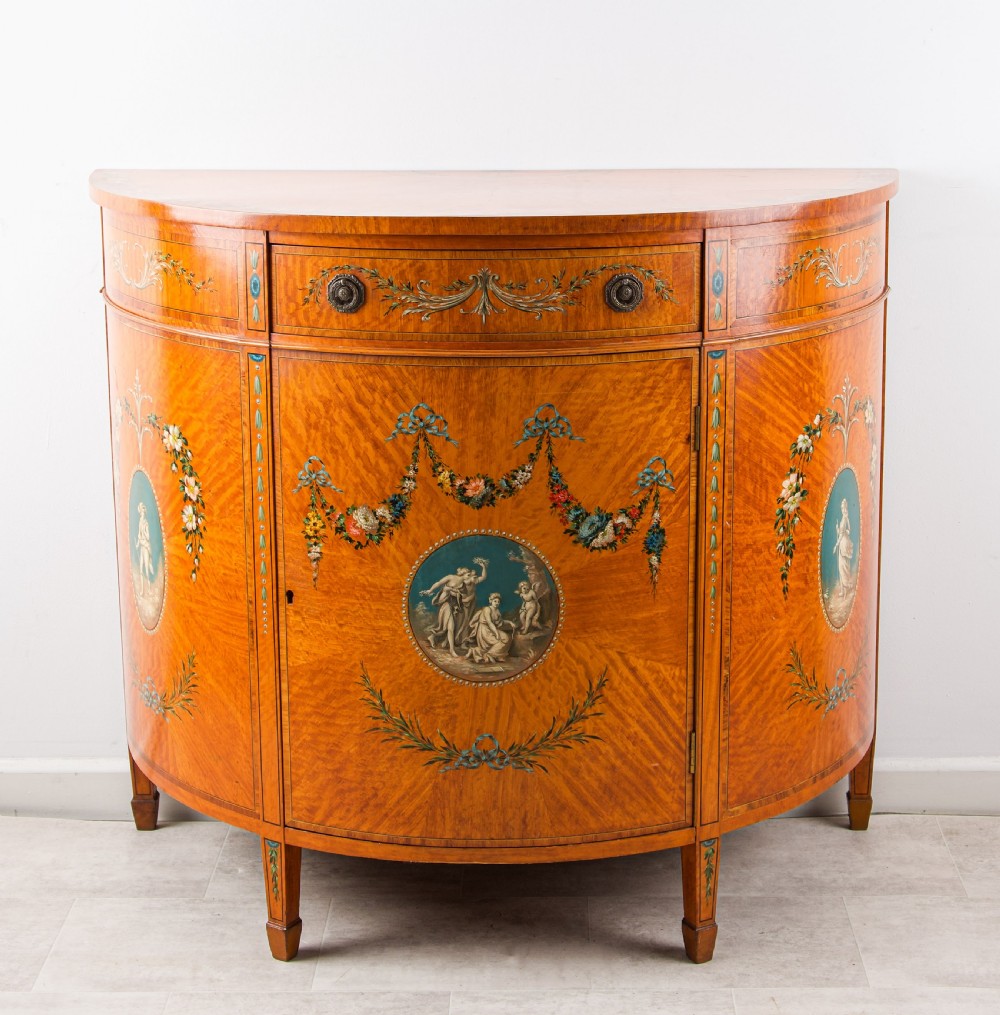 An Edwardian satinwood demi-lune commode with hand-painted classical vignettes, floral garlands, and square tapered legs.
