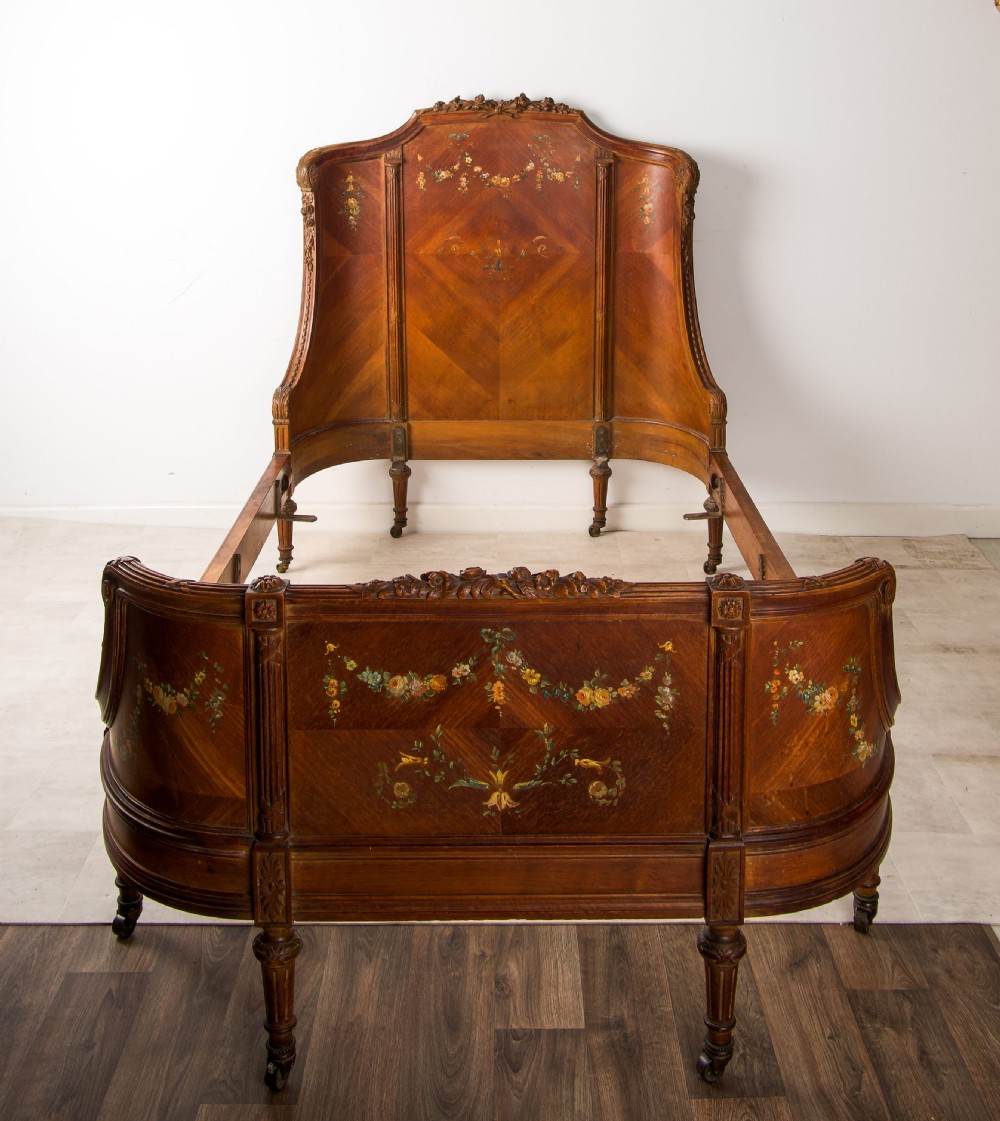 An antique French wooden bed frame featuring a high curved headboard, wrapped footboard, parquetry veneer, and hand-painted floral swags.