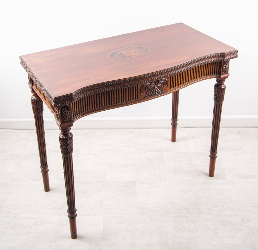 An Edwardian Hepplewhite style mahogany fold-over card table with an oval inlaid central medallion, fluted apron, and tapered fluted legs.