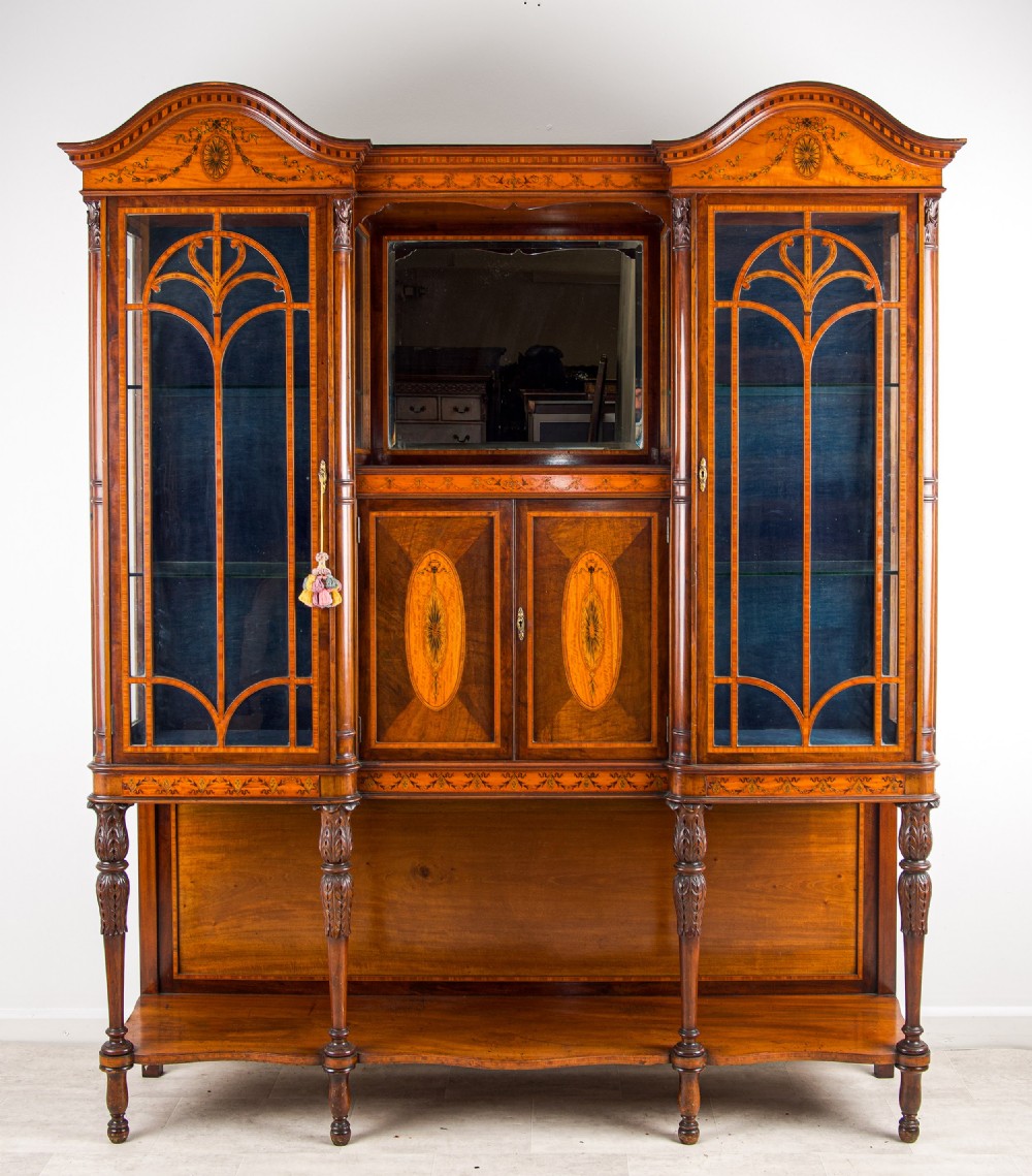 A late Victorian mahogany breakfront display cabinet featuring satinwood marquetry inlay, glazed side doors, a central mirror, and an open lower shelf resting on turned legs.