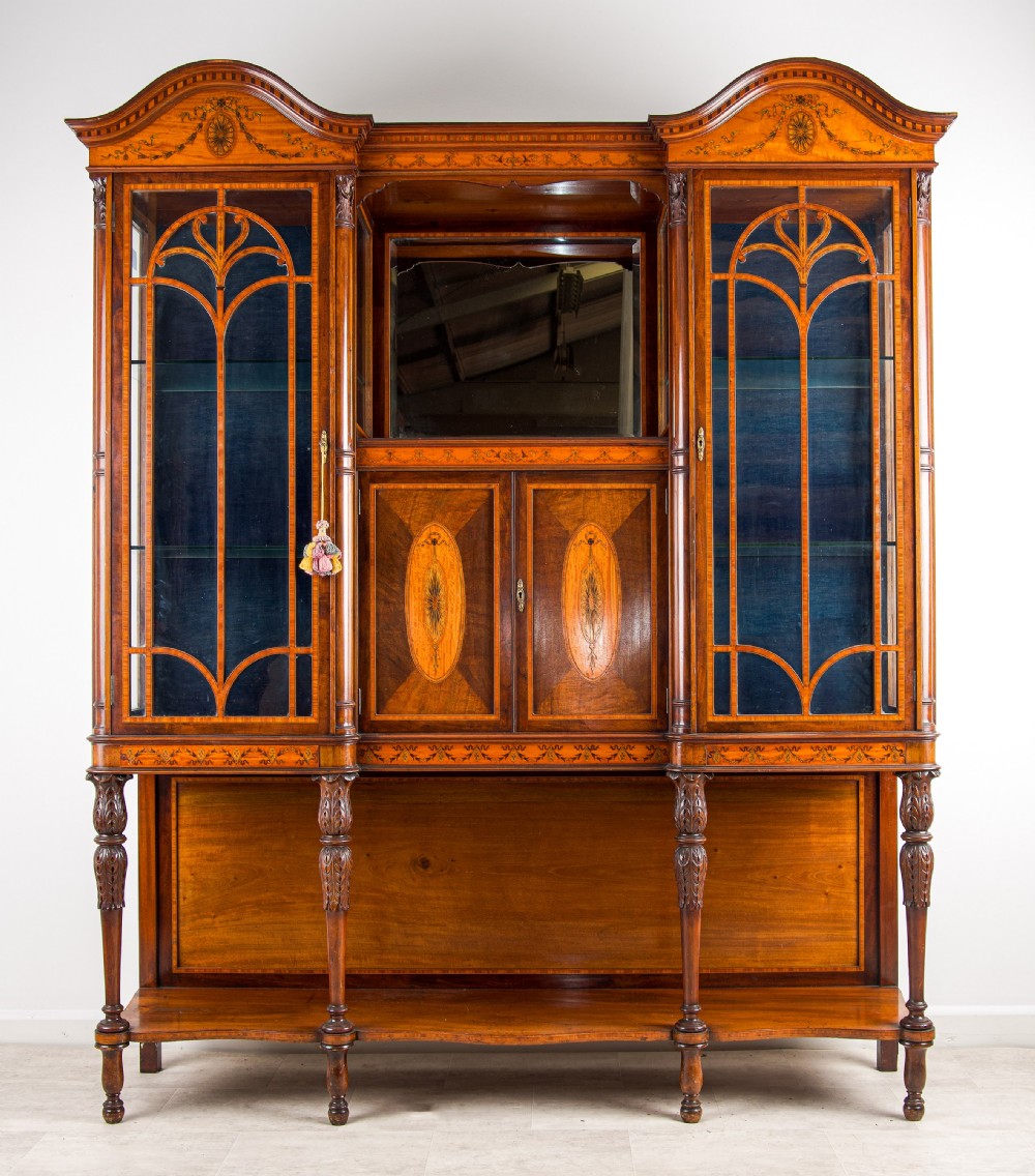 A late Victorian mahogany breakfront display cabinet featuring satinwood marquetry inlay, glazed side doors, a central mirror, and an open lower shelf resting on turned legs. View VI