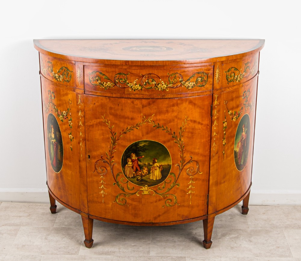 An antique Sheraton Revival semi-circular satinwood side cabinet featuring hand-painted classical figurative ovals, floral swags, and tapered square legs.