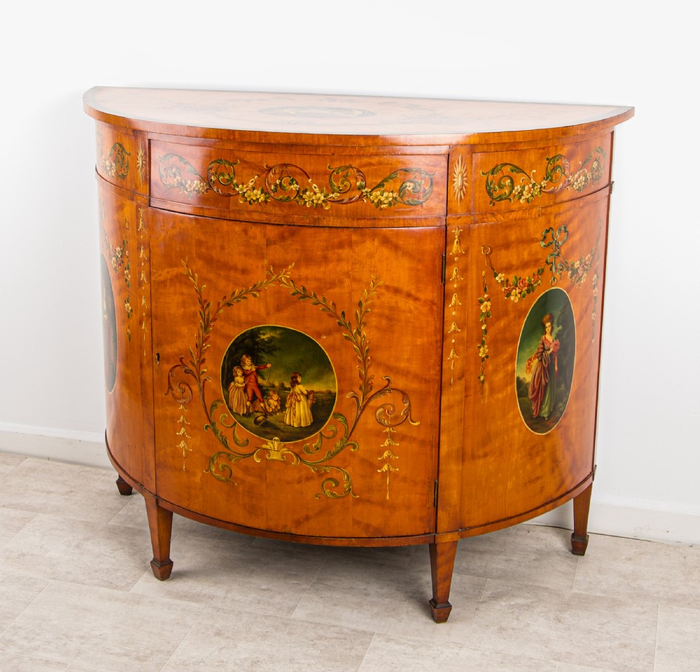 An antique Sheraton Revival semi-circular satinwood side cabinet featuring hand-painted classical figurative ovals, floral swags, and tapered square legs. View II
