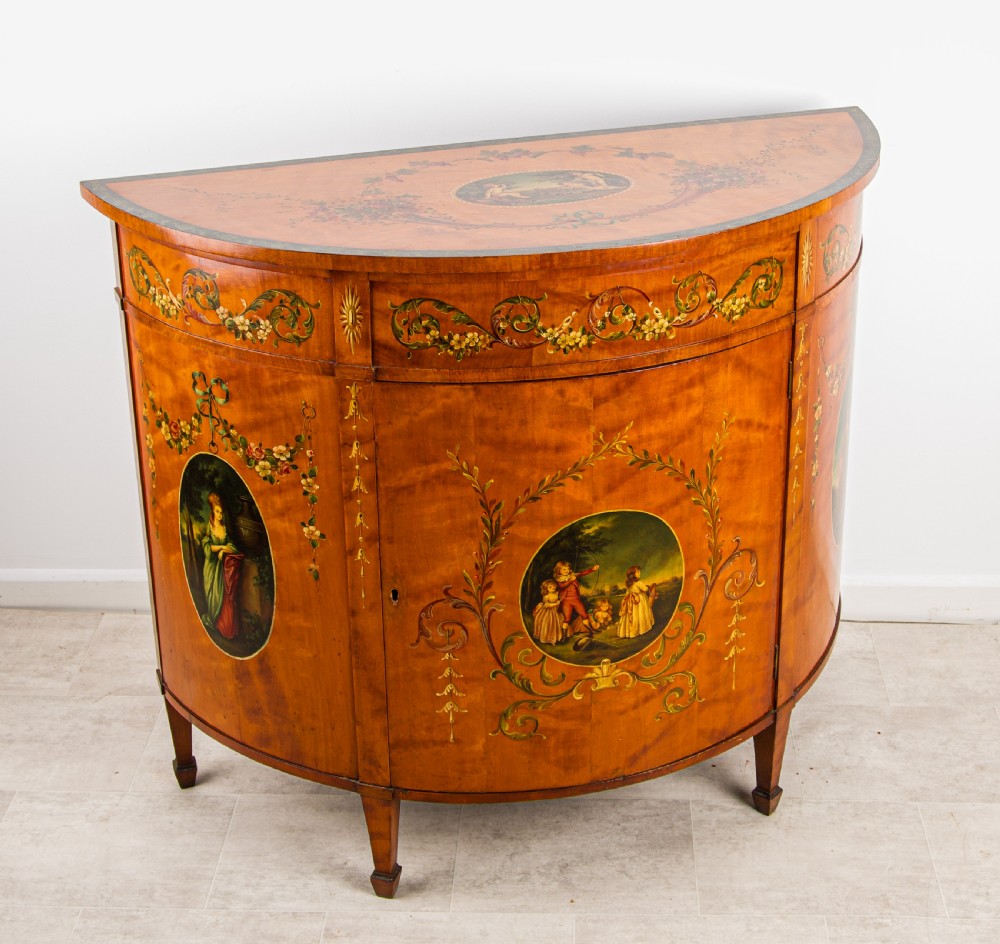An antique Sheraton Revival semi-circular satinwood side cabinet featuring hand-painted classical figurative ovals, floral swags, and tapered square legs. View V