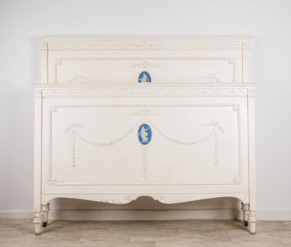 An antique cream-painted French double bed frame featuring carved neoclassical swags, moulding, and central blue Jasperware-style oval plaques.