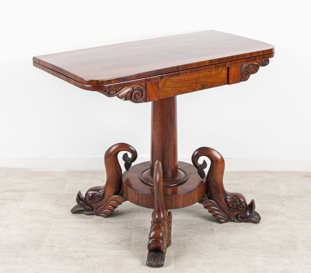 A Regency rosewood games table featuring a swivel fold-over rectangular top set above a central pedestal with four carved dolphin-form feet.