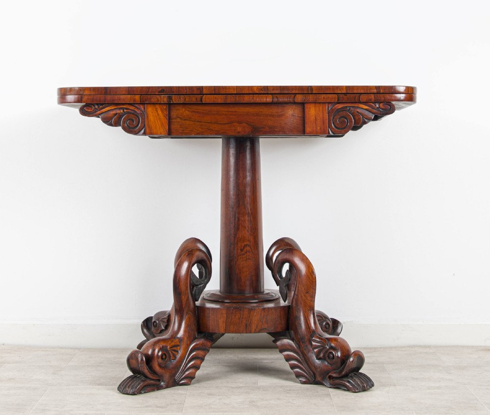 A Regency rosewood games table featuring a swivel fold-over rectangular top set above a central pedestal with four carved dolphin-form feet. View II