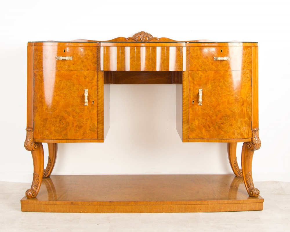 An Art Deco burr walnut sideboard featuring two outer pedestal cupboards, carved cabriole legs, a glass top, and a lower plinth base.