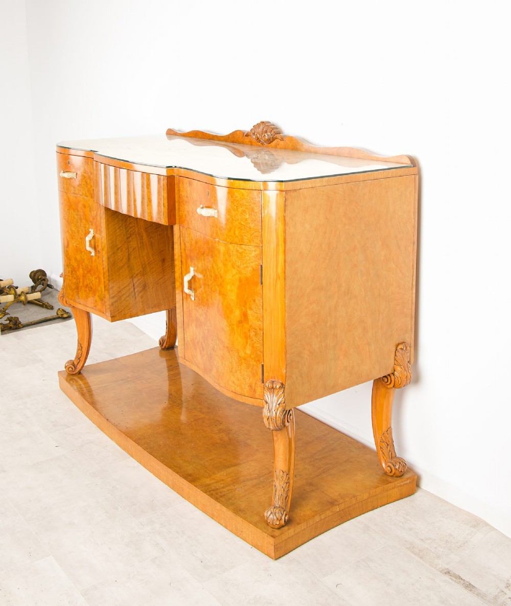 An Art Deco burr walnut sideboard featuring two outer pedestal cupboards, carved cabriole legs, a glass top, and a lower plinth base. View IV