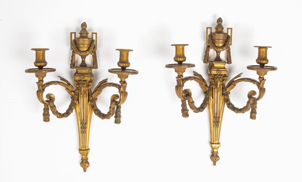 A pair of 19th-century French Neoclassical gilt bronze two-arm wall sconces featuring urn finials, rams head masks, fluted central shafts, and drapery swags.