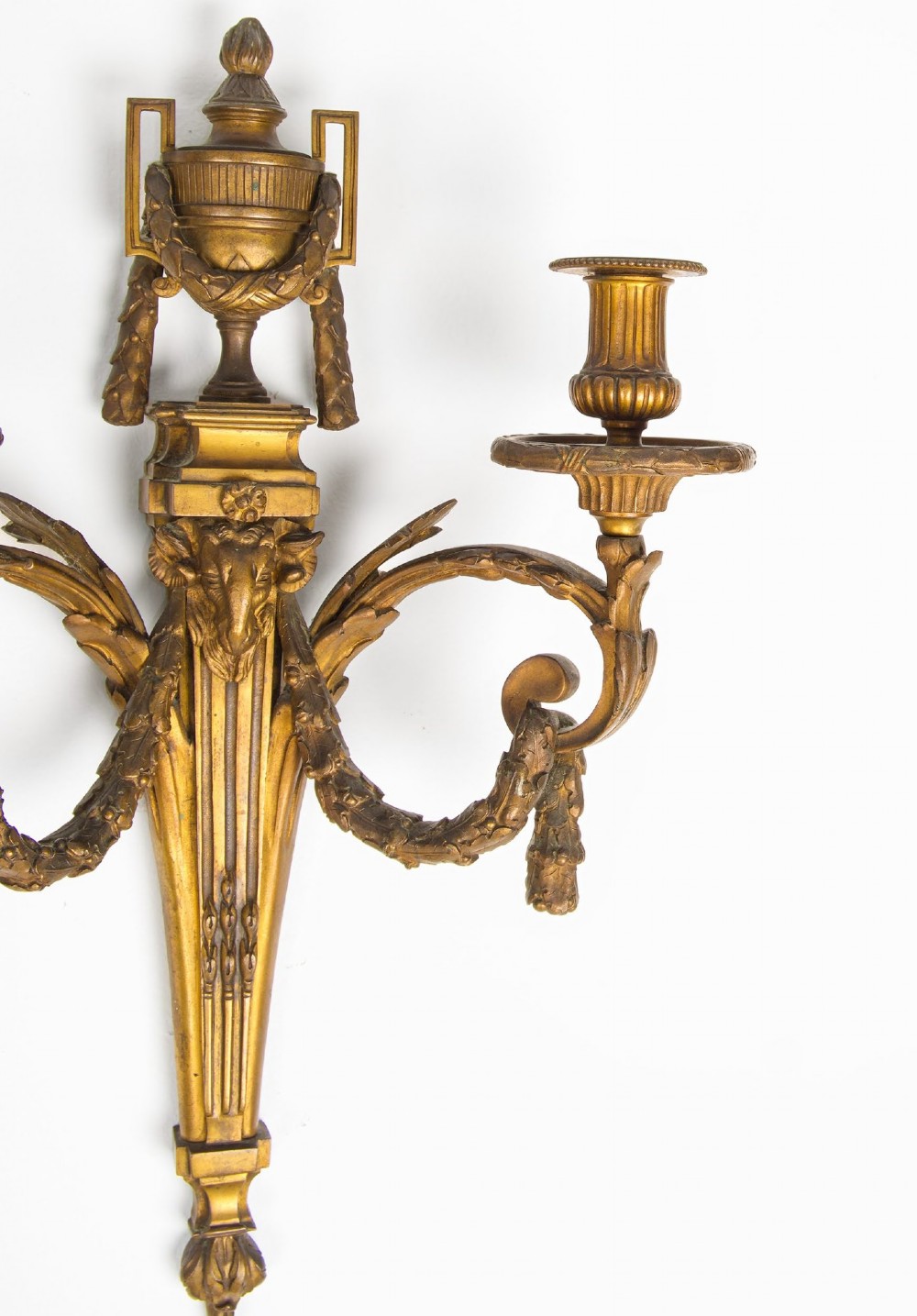 A pair of 19th-century French Neoclassical gilt bronze two-arm wall sconces featuring urn finials, rams head masks, fluted central shafts, and drapery swags. View V