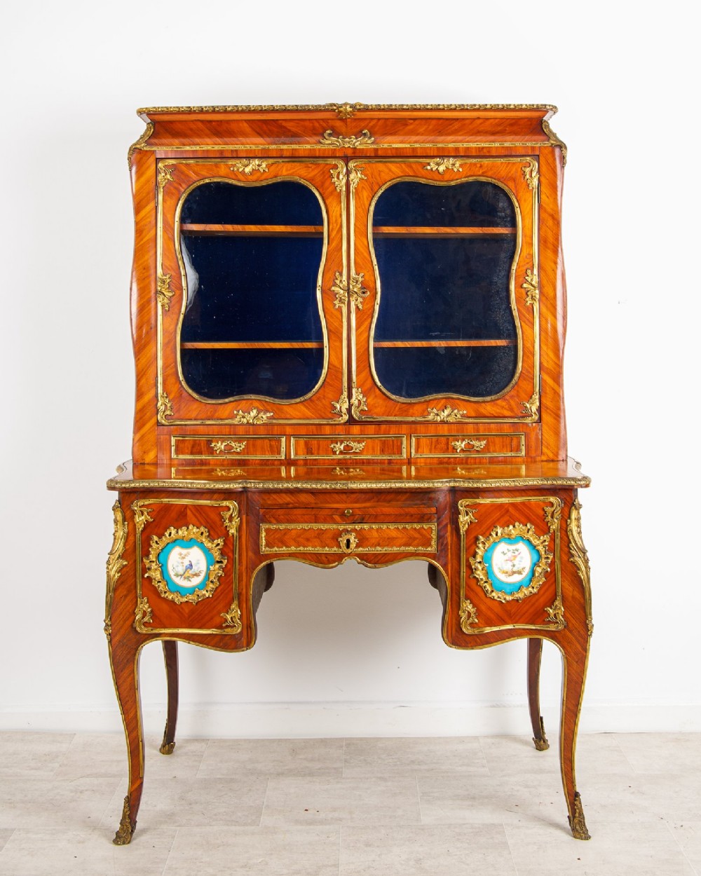 An antique French kingwood Bonheur du Jour cabinet featuring glazed upper doors, ornate ormolu mounts, Sevres style porcelain plaques, and cabriole legs.