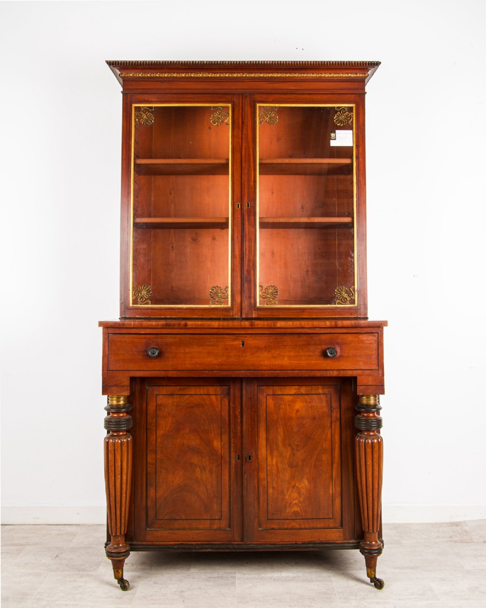 An antique Regency mahogany glazed bookcase cabinet with brass decorative mounts, fluted columns, and a lower two-door cupboard.