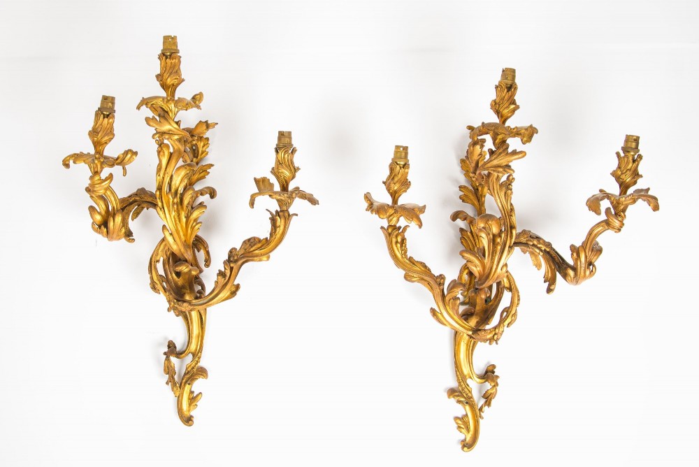 A pair of gilt bronze French ormolu three-branch wall lights decorated with sweeping Rococo acanthus leaf scrollwork.