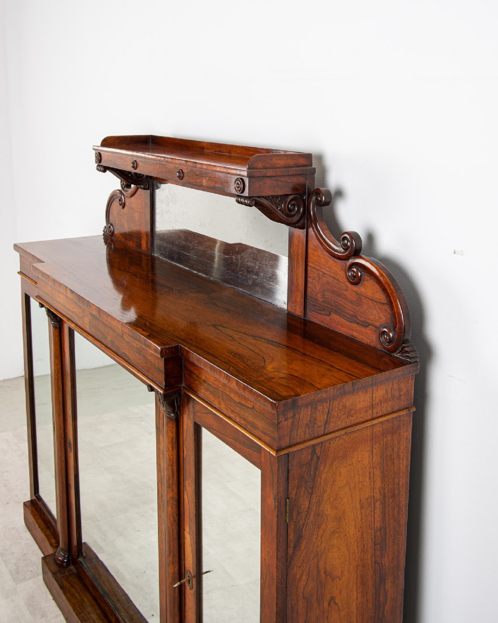 A Victorian rosewood side cabinet featuring a scroll-carved upper back with a shelf and mirror, positioned above three glass-panelled cupboard doors. View IV