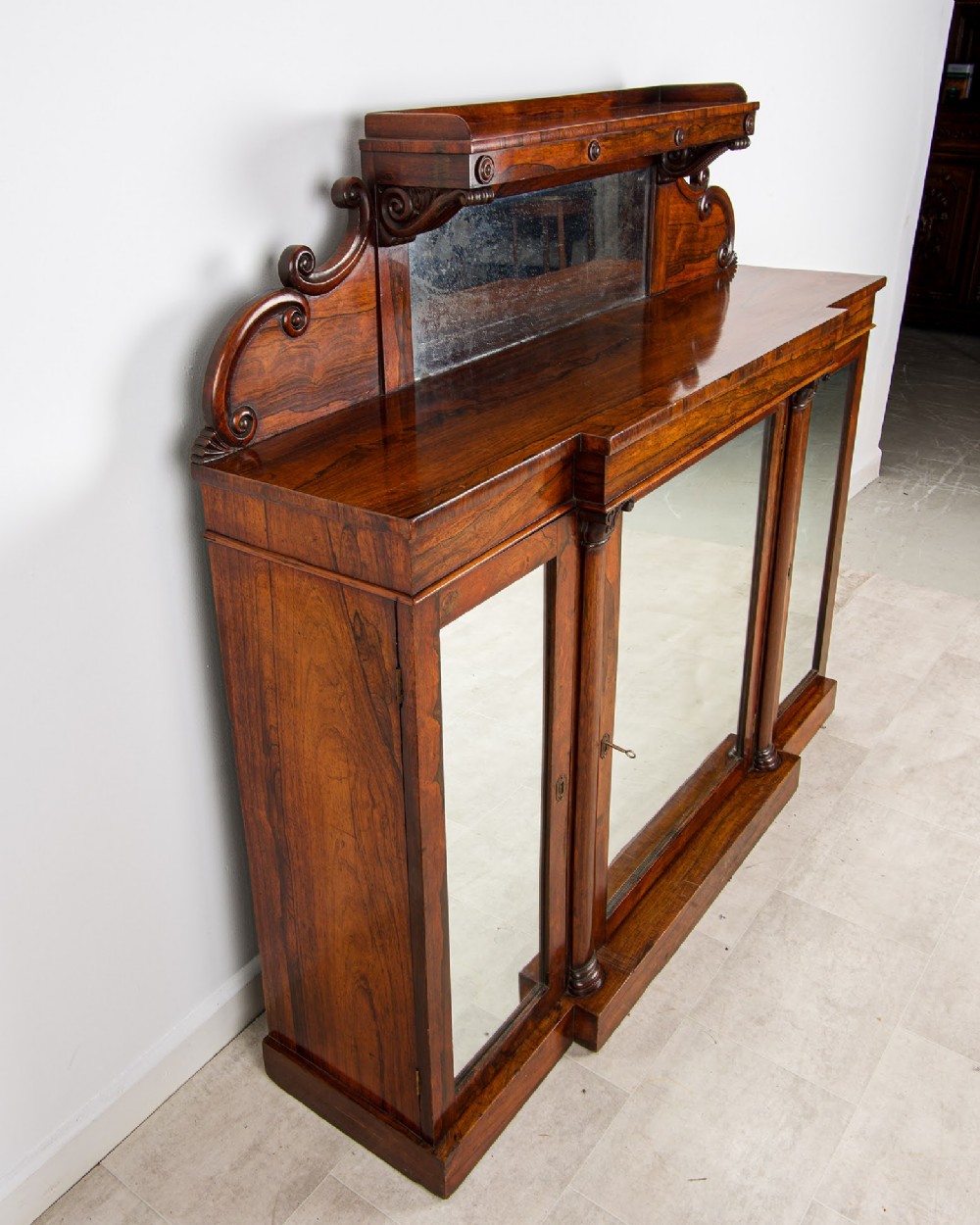 A Victorian rosewood side cabinet featuring a scroll-carved upper back with a shelf and mirror, positioned above three glass-panelled cupboard doors. View VI