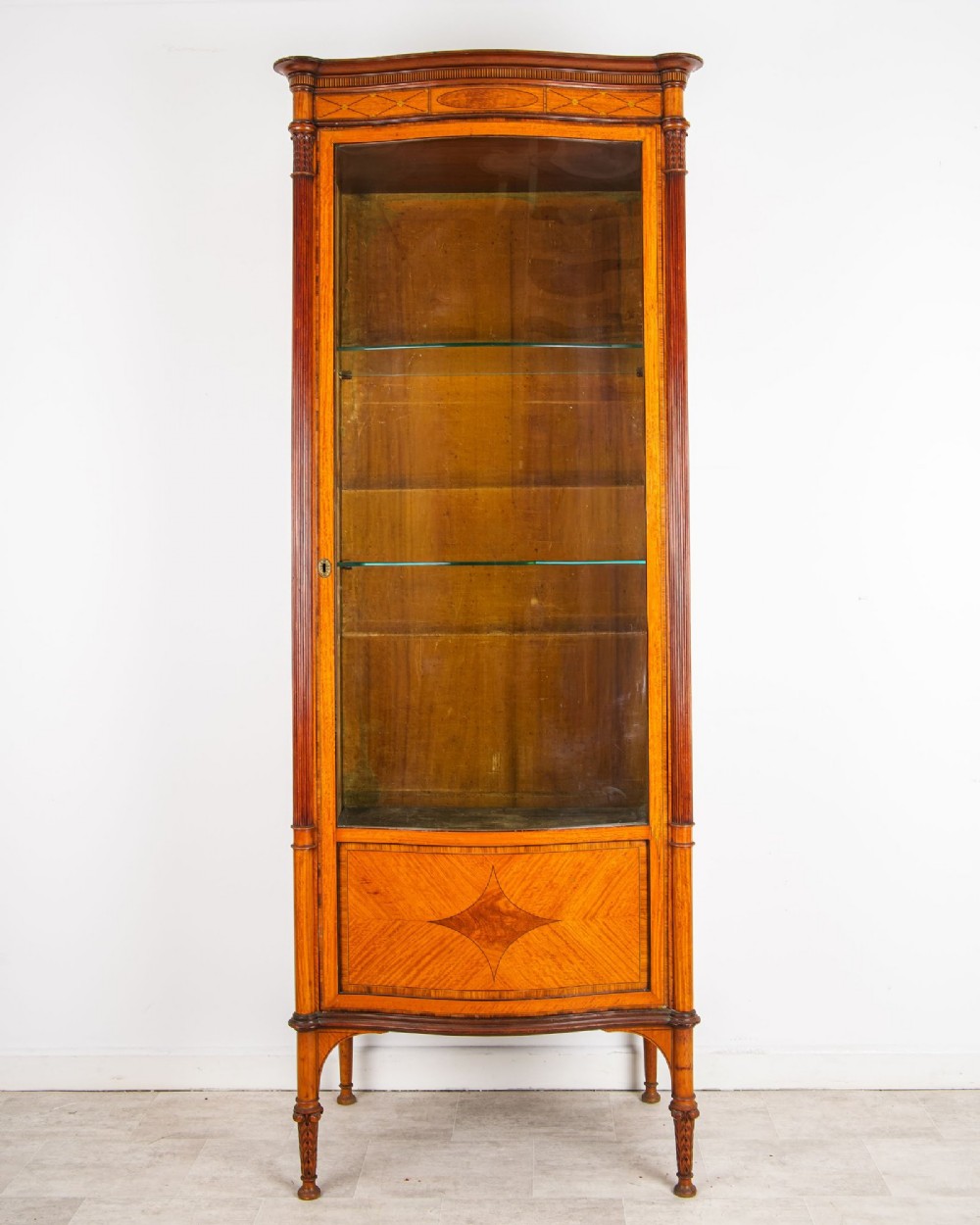 A tall Edwardian satinwood display cabinet featuring a curved glazed door, glass shelves, fluted corner columns, and geometric marquetry inlay.