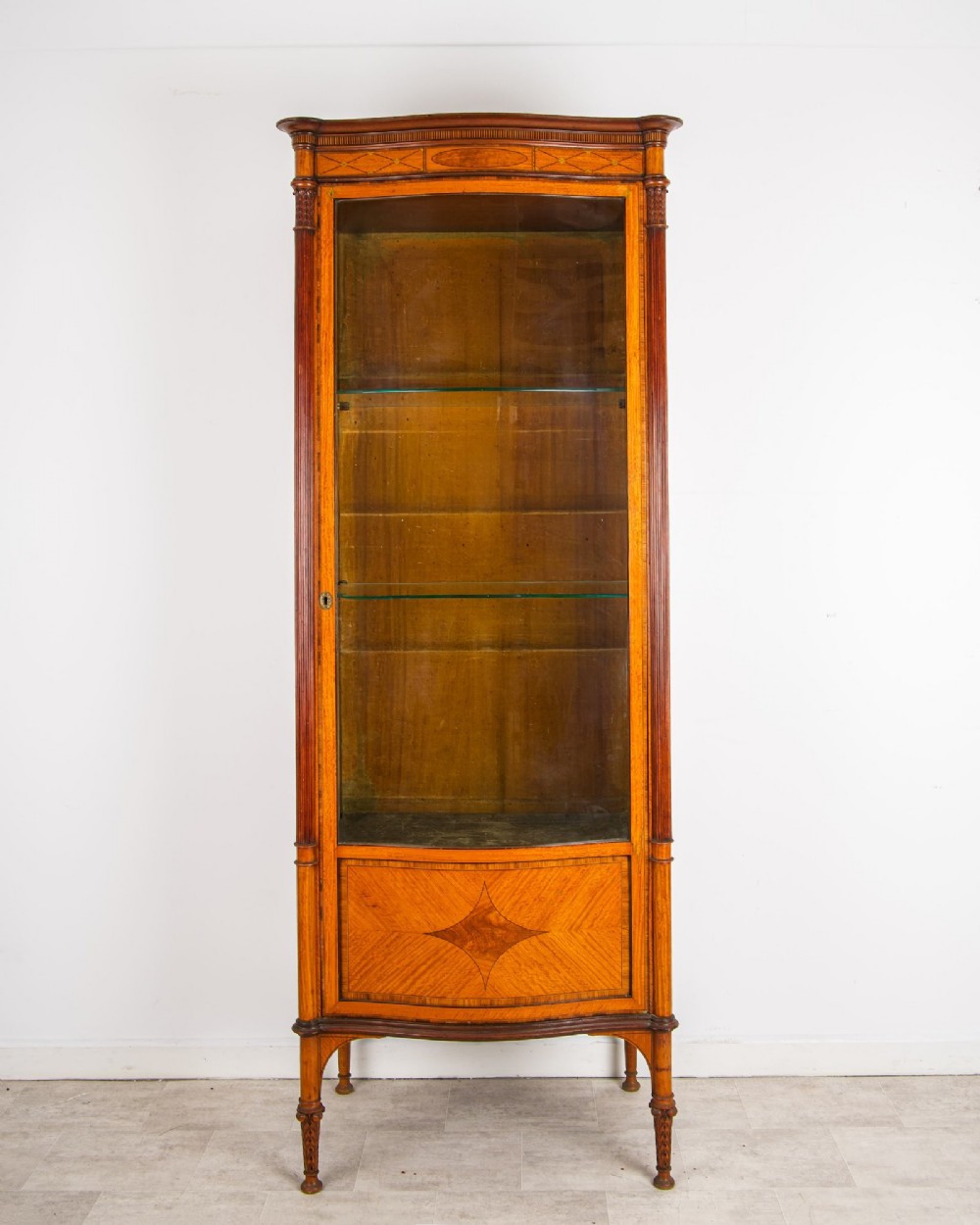 A tall Edwardian satinwood display cabinet featuring a curved glazed door, glass shelves, fluted corner columns, and geometric marquetry inlay. View II